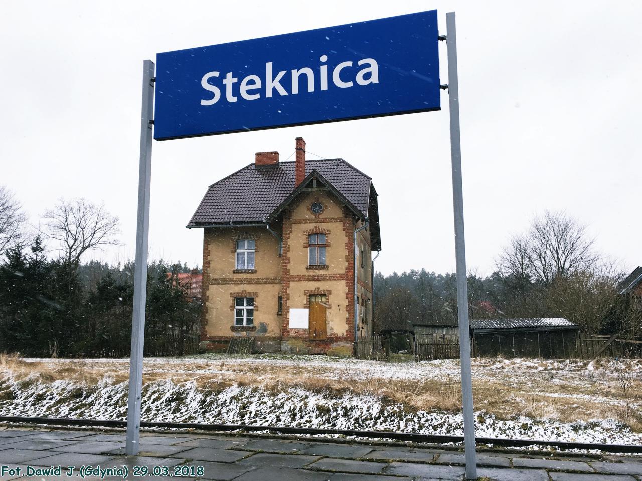 fot. Dawid Jastrzębski 
Kolejowy budynek mieszkalny i pobliski budynek gospodarczy. Obiekty widoczne od strony torów. Tablica z nazwą przystanku osobowego. W lewo Lębork, w prawo Łeba. Obraz