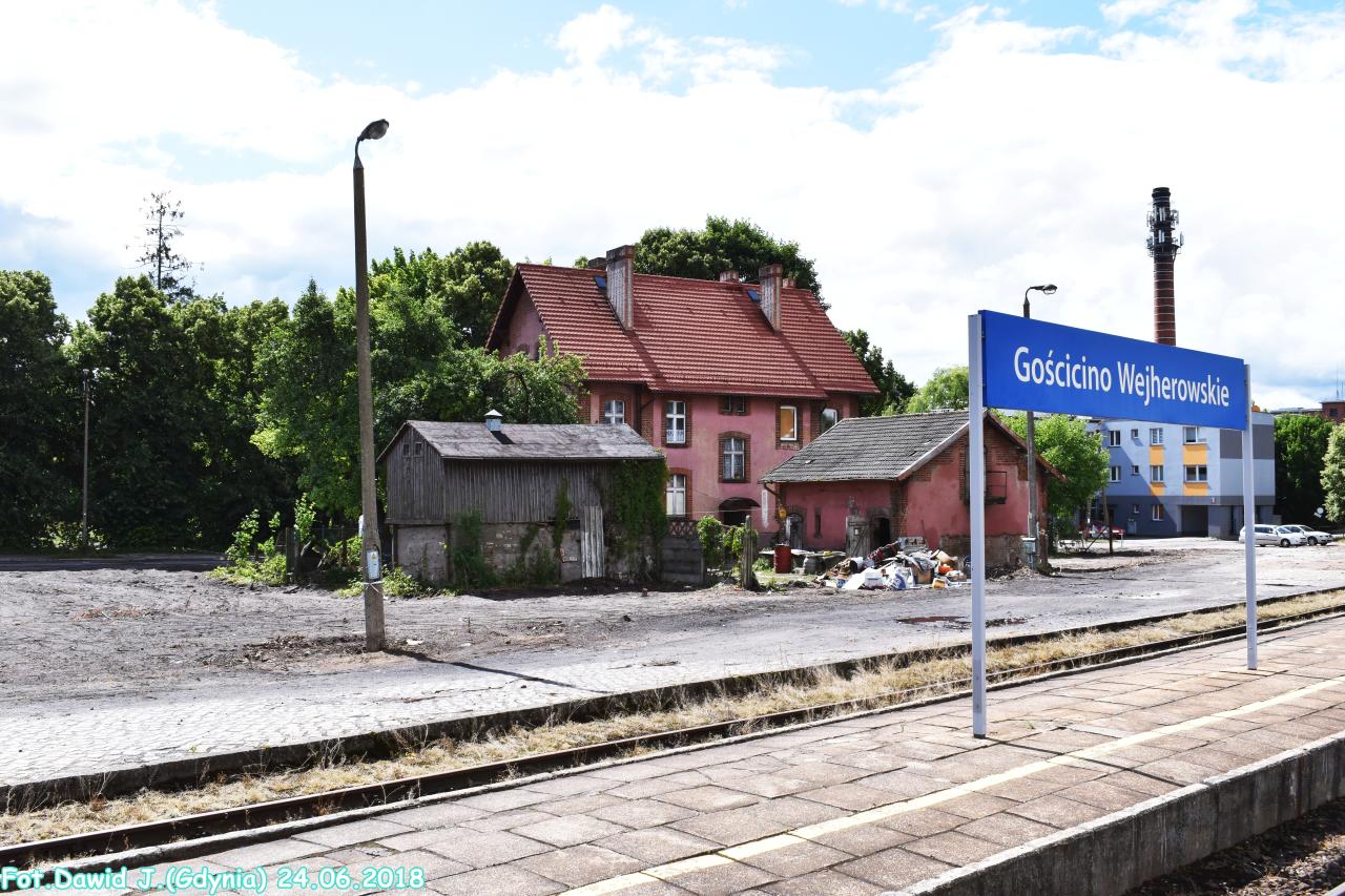 fot. Dawid Jastrzębski 
Kolejowy budynek mieszkalny (ul. Fabryczna 2) i pobliski budynek gospodarczy. Obiekty widoczne od strony torów. Tablica z nazwą stacji. W lewo Wejherowo/Gdynia Główna, w prawo Lębork/Słupsk. Obraz