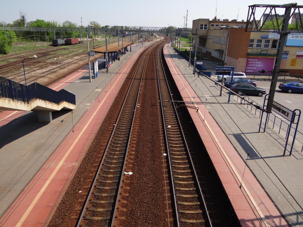fot. Przemysław Więcek 
Po prawej peron pierwszy, boczny. Po lewej peron drugi, wyspowy. Po prawej parking przy ulic Dworcowej na około 50 miejsc. Po prawej dawny budynek dworcowy (ulica Dworcowa 1A). Widok w kierunku Góry Kalwarii i Żyrardowa. 65,8 km/0,1 km. Obraz