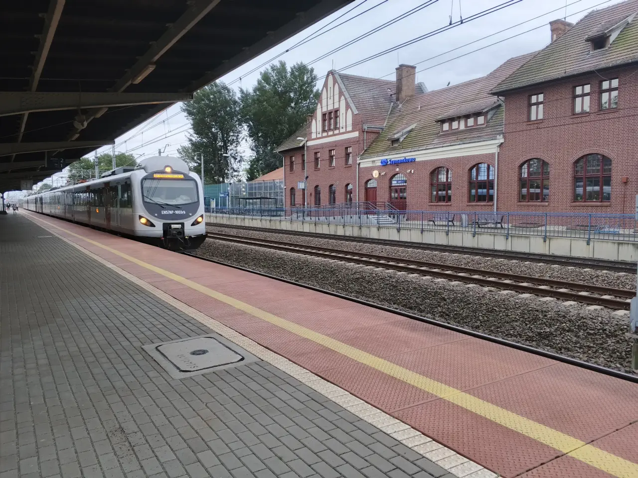 fot. Rafał Wąsowicz 
Szymankowo, budynek dworca od strony peronów oraz EN57AP-1603 wraz z drugą jednostką w obsłudze pociągu osobowego nr 50640/1 do stacji Gdynia Chylonia. Widok w kierunku Malborka oraz Nowego Dworu Gdańskiego. Obraz